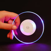 Dessous de verre lumineux tenu de profil dans une main, cercle lumineux bleu-violet allumé sur fond noir.