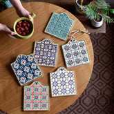 Six dessous de verre en mosaïque aux motifs variés disposés sur une table en bois, avec des mains tenant un bol de fruits rouges.