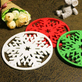 Trois dessous de verre de Noël en forme de flocon (rouge, blanc, vert) posés sur une table en cuir avec un bouquet de roses et un ourson en peluche en arrière-plan