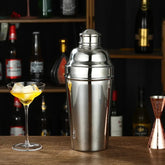 Shaker à cocktail en inox 1L posé sur un comptoir de bar en bois, à côté d'un verre à cocktail garni de glaçons et d'un doseur en cuivre, avec des bouteilles en arrière-plan.