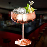 Verre à cocktail coupe en cuivre rempli de glaçons, garni de menthe fraîche, de fleurs et de quartiers de citron vert, sur fond sombre de bar.