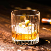 Verre à cocktail Old Fashioned strié rempli de whisky ambré avec glaçon et zeste d'orange, posé sur un comptoir en bois sombre, bouteille floue en arrière-plan