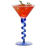 Verre à cocktail à pied torsadé bleu rempli d'un cocktail rouge glacé garni de brins de menthe, sur fond blanc.
