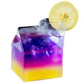 Verre à cocktail en forme de maison sur fond blanc, avec un cocktail au dégradé violet, rose et jaune décoré d'une rondelle de citron.