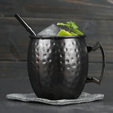 Verre à cocktail Moscow Mule noir garni de glace, de menthe fraîche et d'un quartier de citron vert, avec une paille noire, posé sur un napperon en tissu gris.