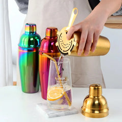 Le set cocktail 4 pièces décliné en trois coloris (arc-en-ciel, rose fuchsia et doré), doseur doré versant une boisson dans un verre.
