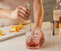 Cuillère à cocktail utilisée pour remuer un cocktail rouge dans un verre, avec zeste d'orange et bouteilles d'alcool en arrière-plan