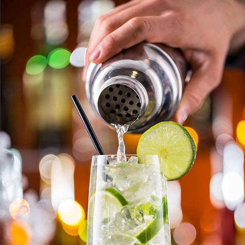 Gros plan sur le shaker versant un cocktail dans un verre garni d'une rondelle de citron vert, avec les lumières du bar en arrière-plan flou.