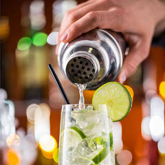 Gros plan sur le shaker versant un cocktail dans un verre garni d'une rondelle de citron vert, avec les lumières du bar en arrière-plan flou.