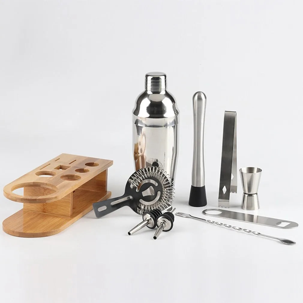 Vue éclatée de tous les accessoires du kit cocktail — shaker, pilon, pince, doseur, passoire, cuillère et bouchons verseurs — posés à côté de leur support en bambou vide.