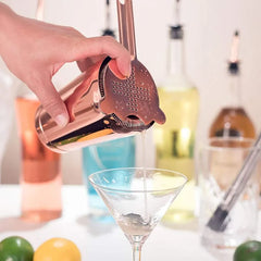 Gros plan sur la passoire Hawthorne rose gold du kit versant un cocktail dans un verre à martini