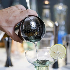 Gros plan sur le shaker Boston versant un cocktail filtré dans une coupe garnie d'une rondelle de citron