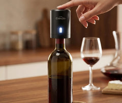 Main appuyant sur le bouton du bouchon pompe à air fixé sur une bouteille de vin ouverte, posée sur une table en bois avec un verre de vin rouge et du fromage en arrière-plan.