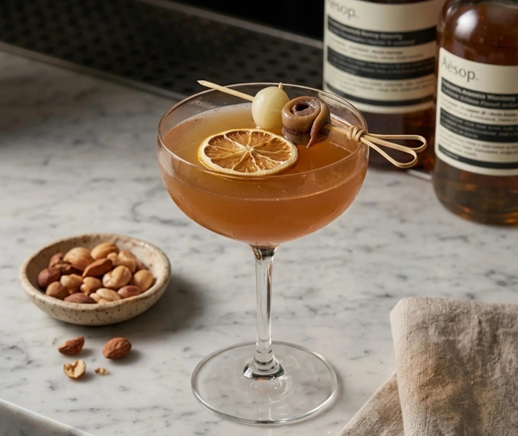 Cocktail ambré garni d'une rondelle d'agrume séchée, d'un cornichon et d'un pic à cocktail en bambou, posé sur un bar en marbre avec bouteilles et bol de fruits secs en arrière-plan.