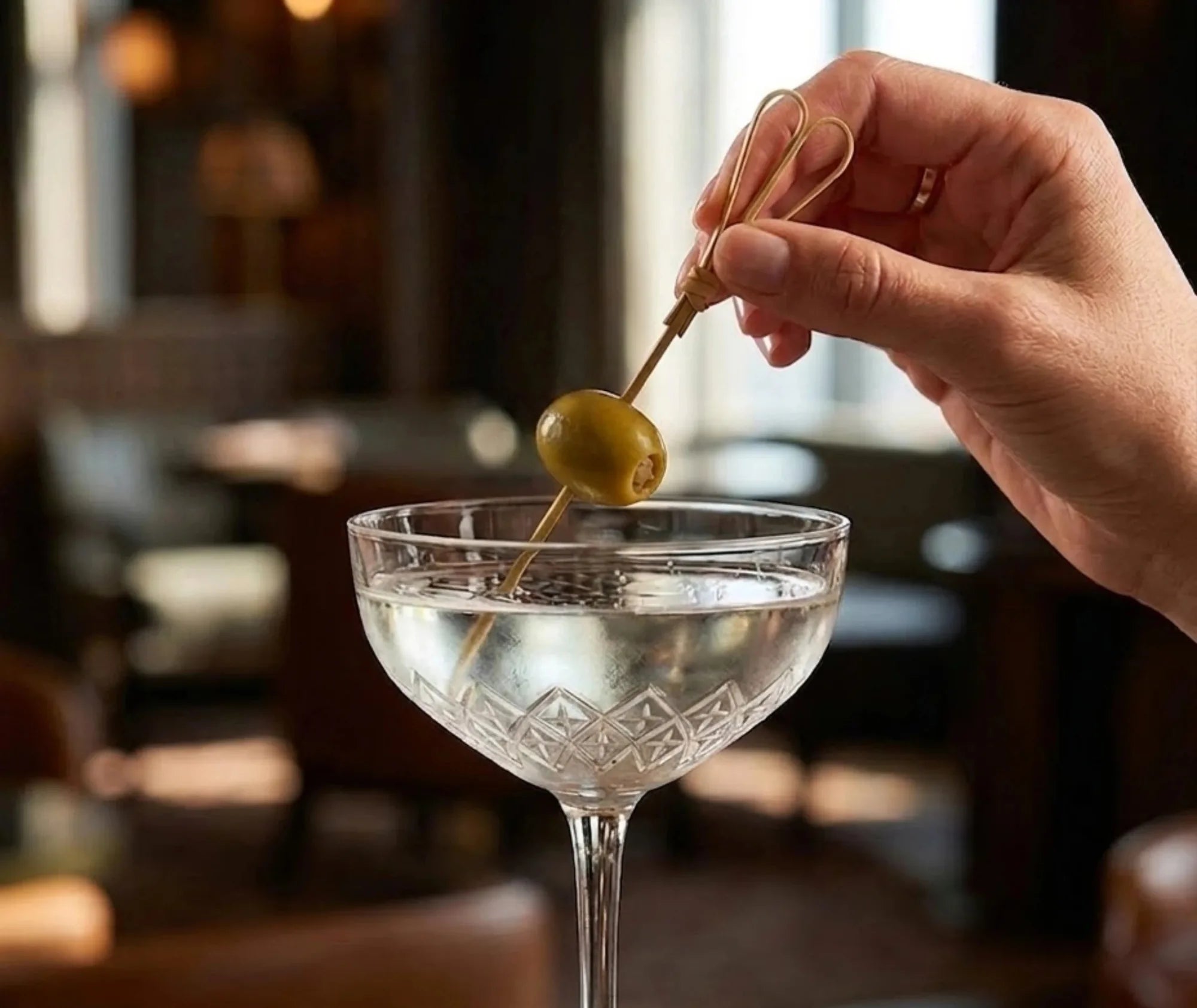 Main tenant un pic à cocktail doré piqué d'une olive verte, au-dessus d'un verre à martini transparent.