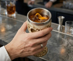Main tenant le calice tête de mort rempli d'un cocktail ambré avec glaçons et zeste d'orange au bar