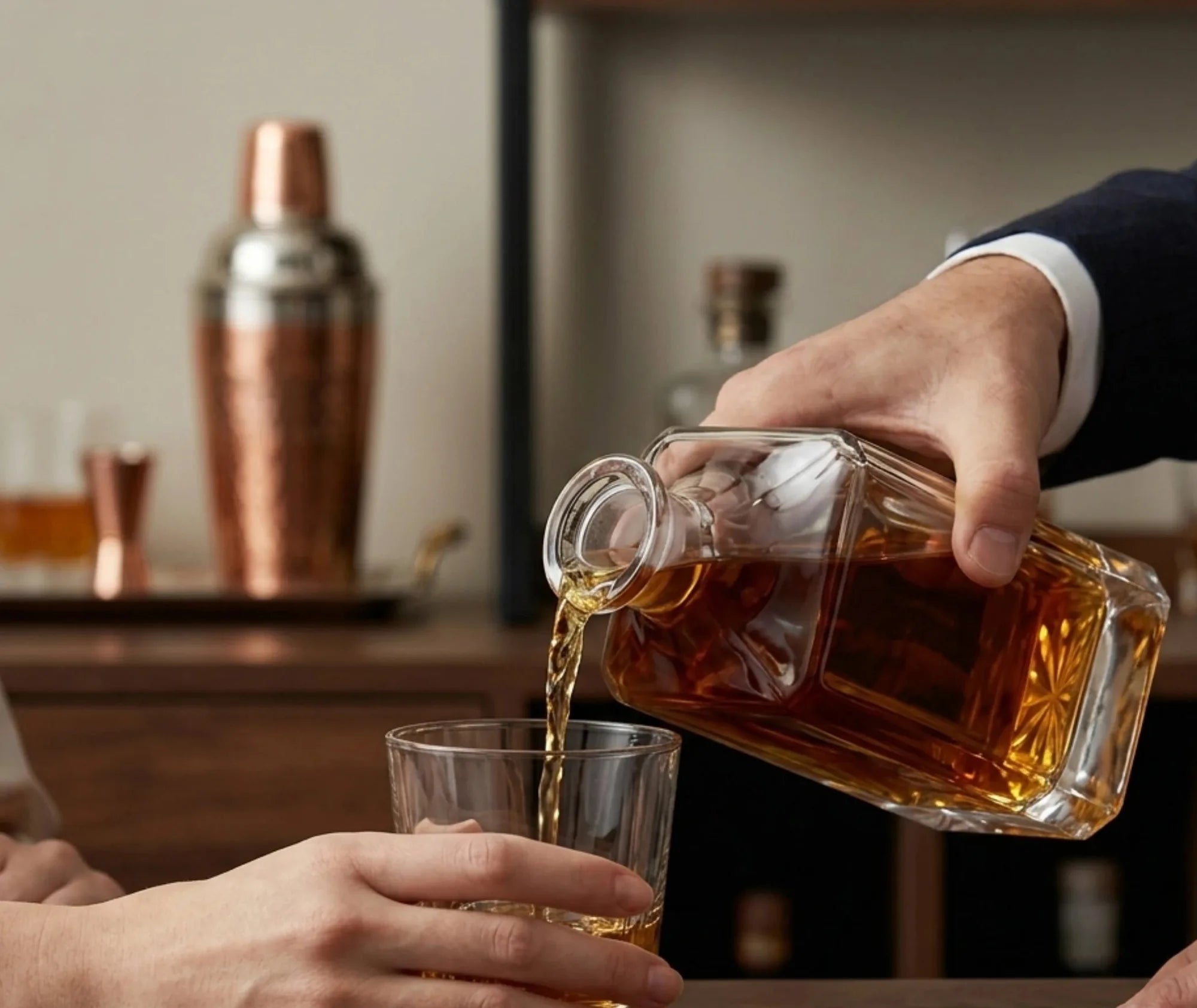 Un barman en costume verse du whisky depuis la carafe carrée dans un verre, avec un shaker en cuivre en arrière-plan.