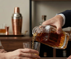 Un barman en costume verse du whisky depuis la carafe carrée dans un verre, avec un shaker en cuivre en arrière-plan.