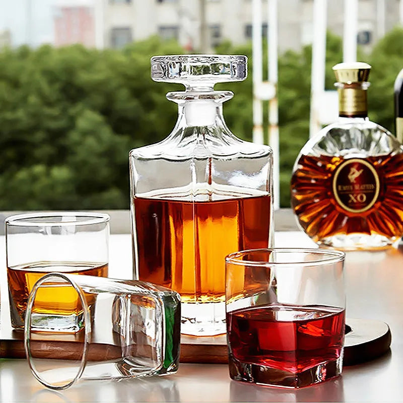 La carafe à whisky carrée posée sur une table extérieure entourée de verres à whisky et d'une bouteille de cognac XO, avec de la verdure en arrière-plan.