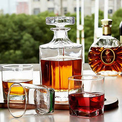 La carafe à whisky carrée posée sur une table extérieure entourée de verres à whisky et d'une bouteille de cognac XO, avec de la verdure en arrière-plan.