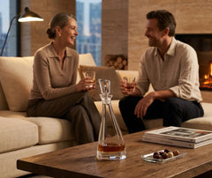 Carafe à whisky moderne posée sur une table basse en bois devant un couple souriant installé dans un salon cosy avec cheminée.