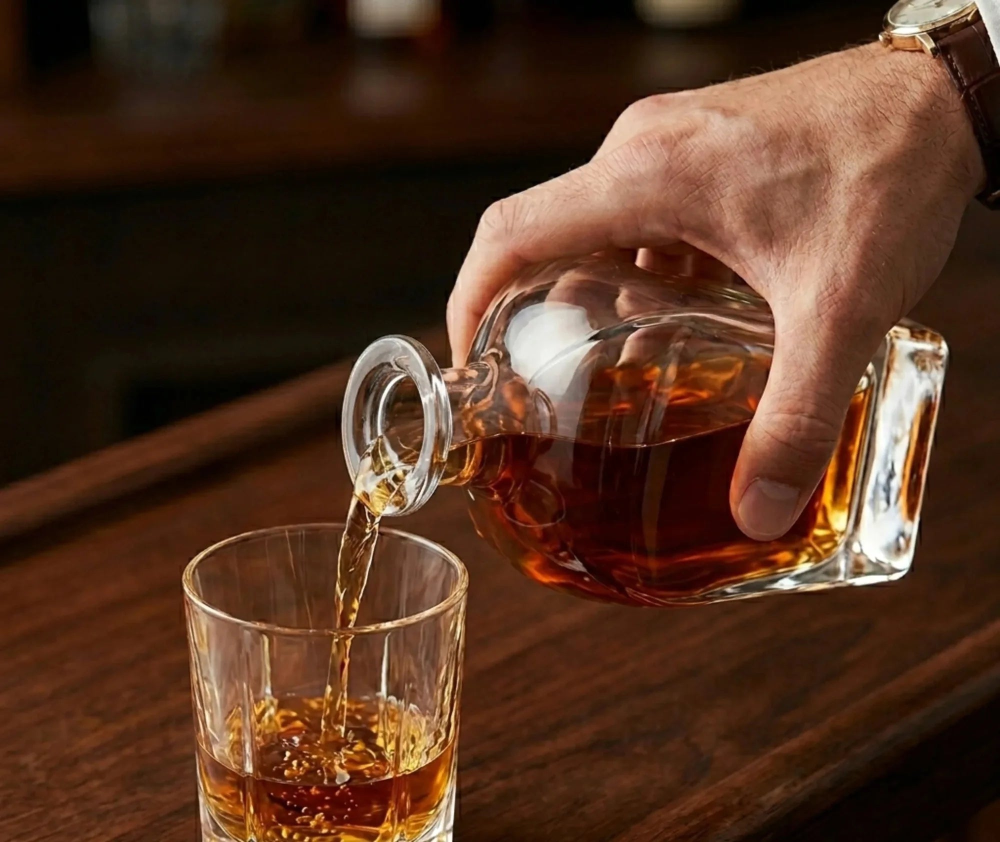 Une main verse du whisky depuis la carafe dans un verre, sur un comptoir de bar en bois sombre.