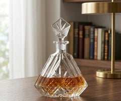 Carafe à whisky en cristal remplie posée sur un bureau en bois, avec rideaux blancs et bibliothèque en arrière-plan.