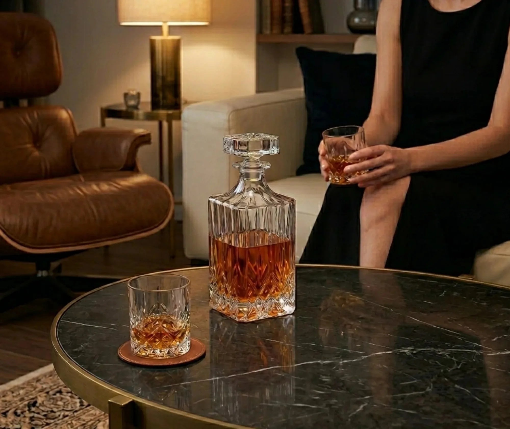 Carafe à whisky en verre taillé posée sur une table basse en marbre dans un salon cosy, avec deux verres de whisky et une femme assise en arrière-plan.