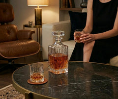 Carafe à whisky en verre taillé posée sur une table basse en marbre dans un salon cosy, avec deux verres de whisky et une femme assise en arrière-plan.