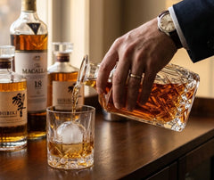 Un homme en costume verse du whisky depuis la carafe en verre taillé dans un verre à glaçon, avec des bouteilles de whisky haut de gamme en arrière-plan.