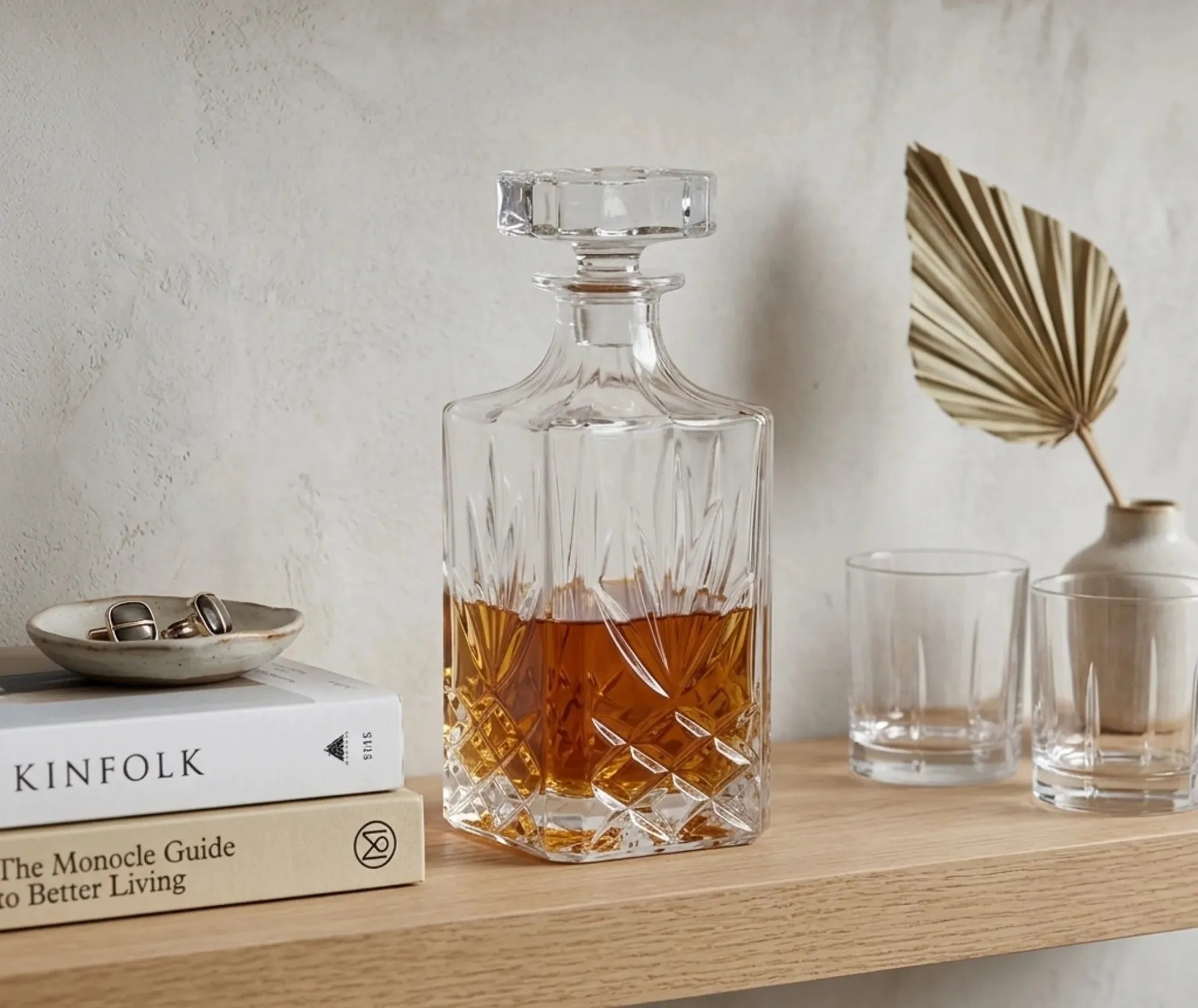 Carafe à whisky sans plomb remplie posée sur une étagère en bois décorée de livres, verres et feuille de palmier séchée.
