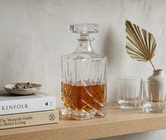 Carafe à whisky sans plomb remplie posée sur une étagère en bois décorée de livres, verres et feuille de palmier séchée.