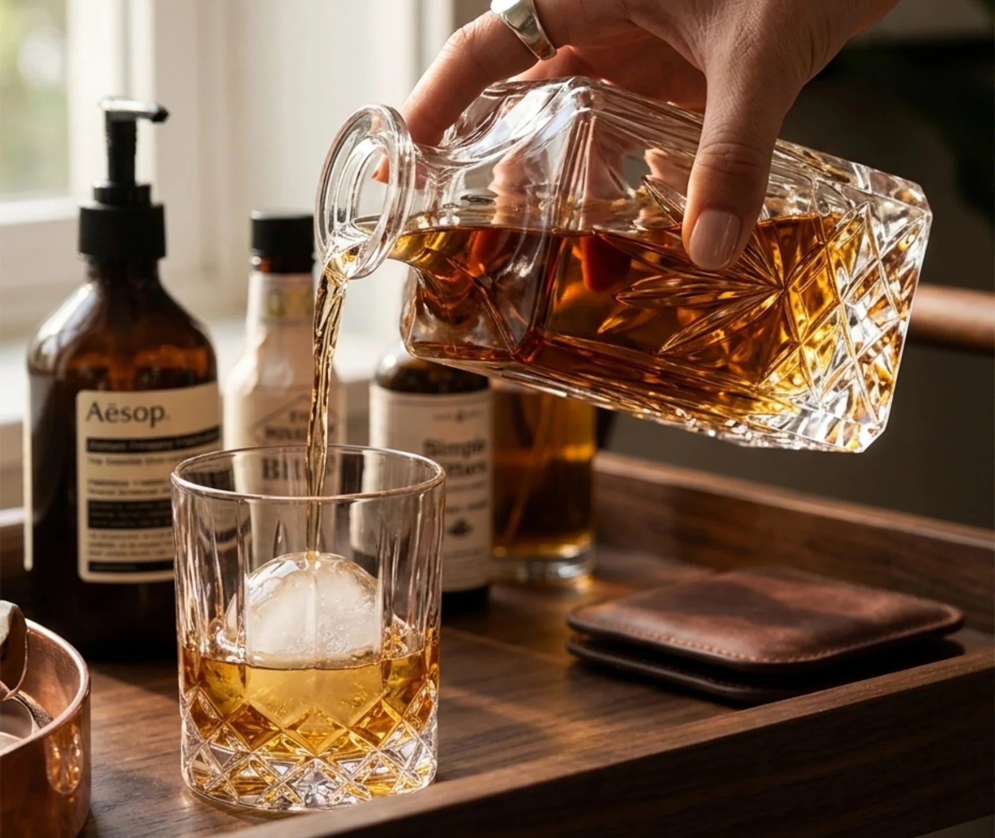 Main versant du whisky depuis la carafe sans plomb dans un verre à glaçon, sur un plateau en bois avec accessoires de bar.