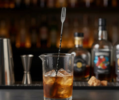 Cuillère de mélange plantée dans un verre de cocktail ambré rempli de glaçons, devant des bouteilles de spiritueux floues au bar.