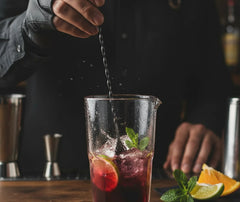 Barman versant un cocktail rouge à la cuillère de mélange dans un verre à mélange garni de glaçons, citron vert et menthe.