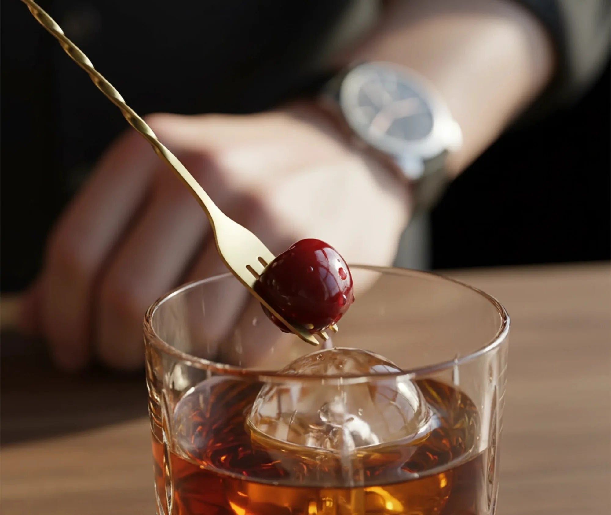 Gros plan de la fourchette dorée piquant une cerise confite au-dessus d'un verre à cocktail avec glaçon sphérique