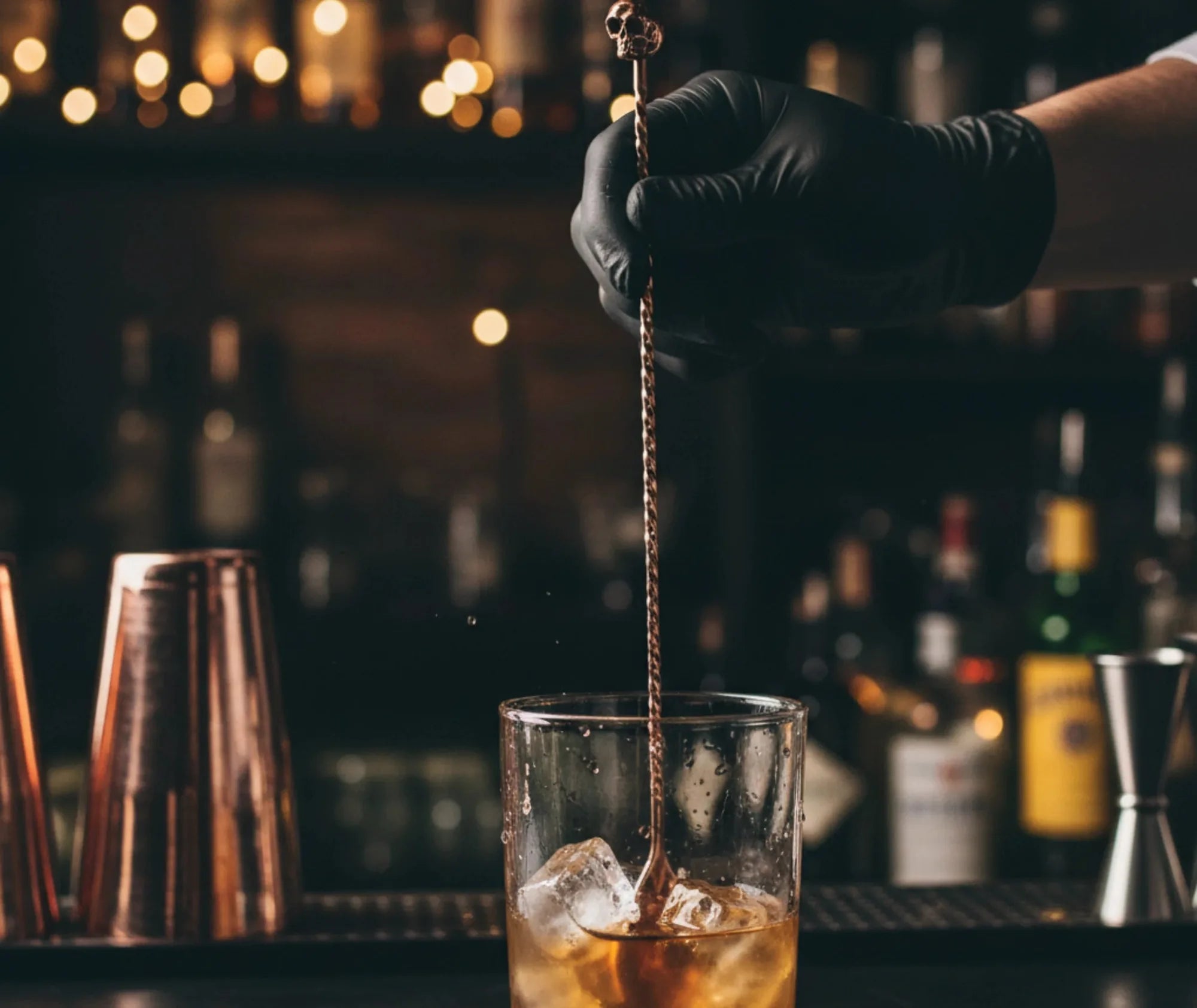 Un barman verse un cocktail ambré à l'aide de la cuillère à cocktail tête de mort dans une ambiance de bar sombre.