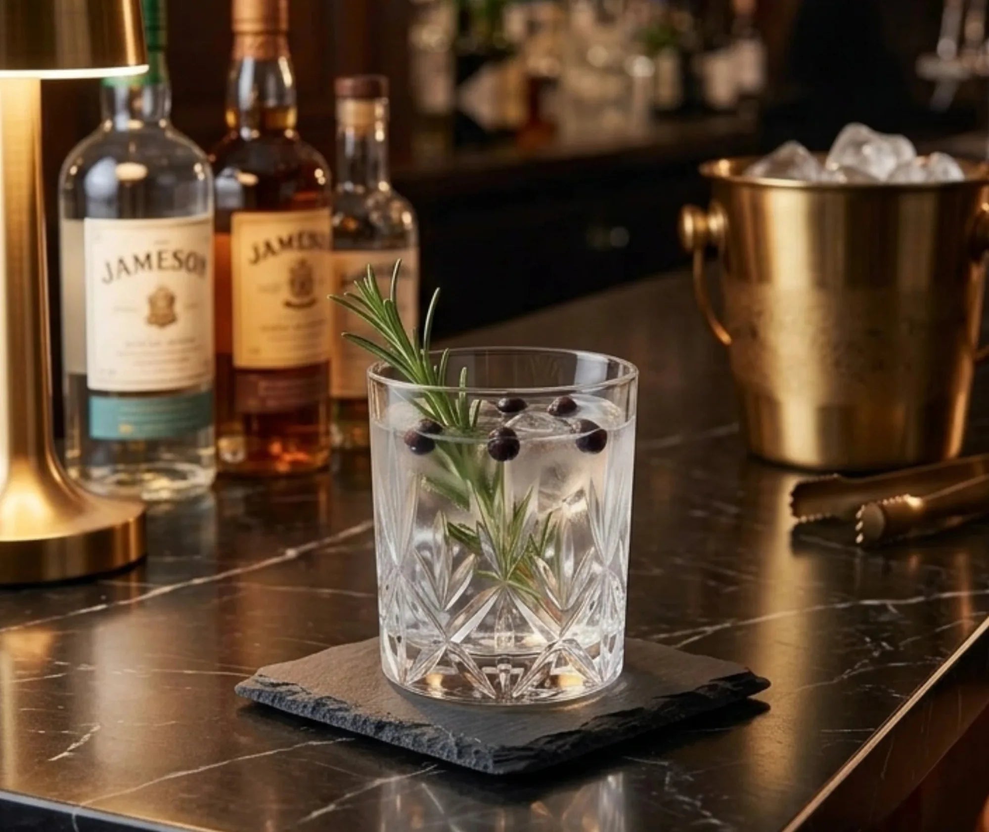 Dessous de verre en ardoise sous un verre à cocktail garni de romarin et de baies de genièvre dans un bar