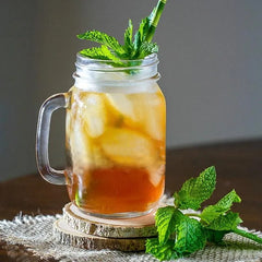 Verre style bocal rempli de thé glacé à la menthe posé sur un dessous de verre en bois, entouré de feuilles de menthe fraîche.