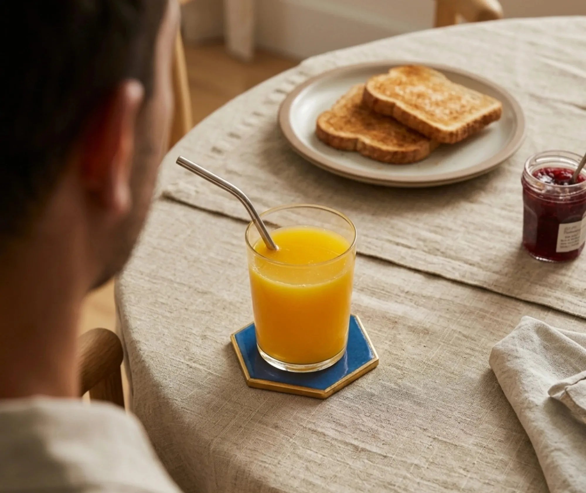 Verre de jus d'orange avec paille posé sur un dessous de verre bleu, à côté d'une assiette de tartines grillées et d'un pot de confiture, lors d'un petit-déjeuner.