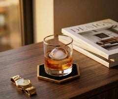 Verre de whisky avec glaçon rond posé sur un dessous de verre noir et doré, sur un bureau en bois avec une montre et un magazine déco.