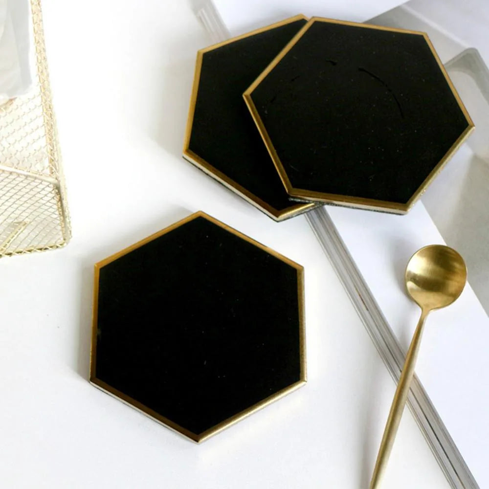 Trois dessous de verre hexagonaux noirs disposés sur une table blanche avec une corbeille dorée, une cuillère et un magazine.