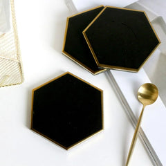 Trois dessous de verre hexagonaux noirs disposés sur une table blanche avec une corbeille dorée, une cuillère et un magazine.
