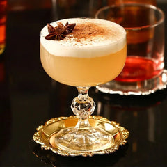 Cocktail doré garni d'anis étoilé et de poudre d'épices, servi dans une coupe posée sur un dessous de verre doré.