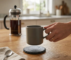 Dessous de verre en ardoise posé sur un plan de travail en bois, une main y dépose une tasse de café dans une cuisine