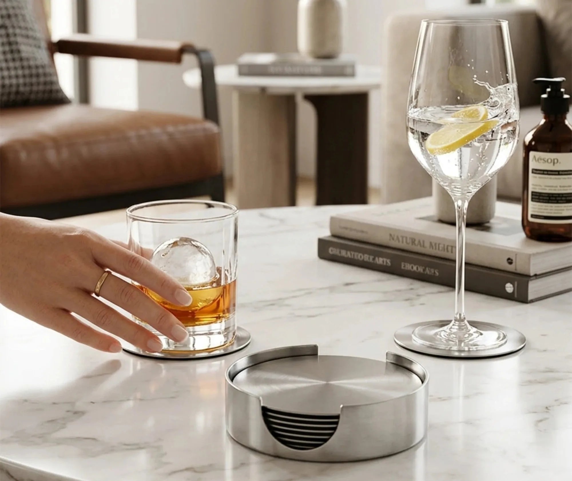 Une main attrape un verre de whisky posé sur un dessous de verre en inox, à côté d'une coupe de vin sur son propre dessous de verre et du boîtier de rangement, sur une table en marbre.