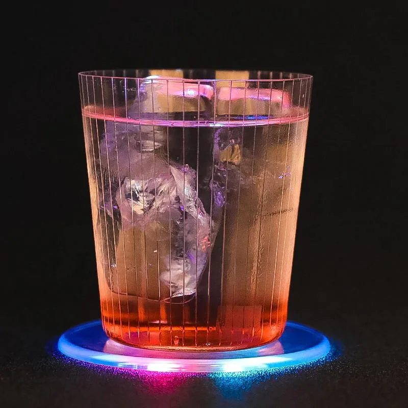 Verre de cocktail rosé avec glaçon posé sur un dessous de verre lumineux aux teintes bleu et rose, fond noir.