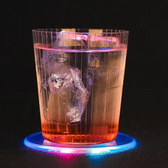 Verre de cocktail rosé avec glaçon posé sur un dessous de verre lumineux aux teintes bleu et rose, fond noir.
