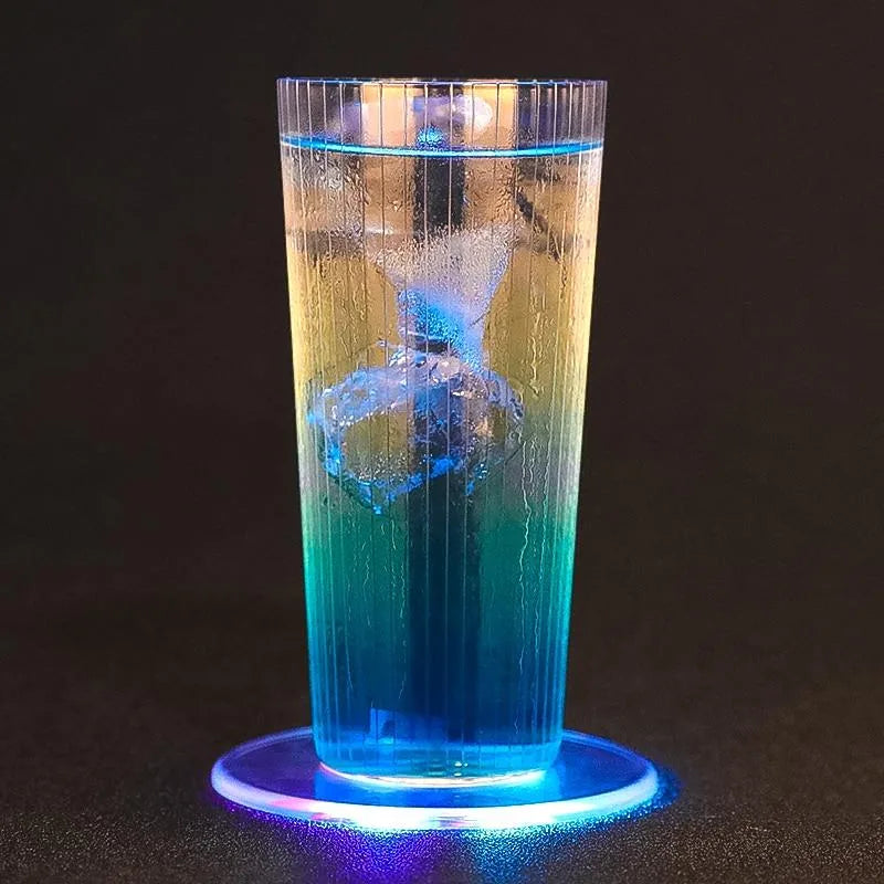 Long drink au dégradé jaune-vert-bleu avec glaçon, posé sur un dessous de verre lumineux bleu sur fond noir.