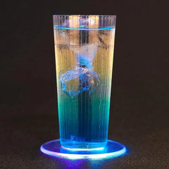 Long drink au dégradé jaune-vert-bleu avec glaçon, posé sur un dessous de verre lumineux bleu sur fond noir.